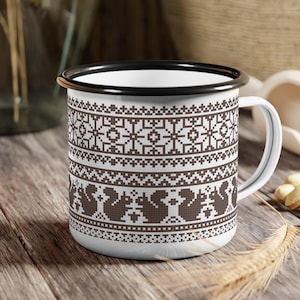 May include: White enamel mug with a black rim and handle. The mug features a brown and white geometric pattern with squirrels. The mug is sitting on a wooden surface.