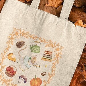 May include: Cream-colored tote bag with a whimsical illustration of a mouse, teapot, books, and pumpkin. The design is framed by a floral border. The bag rests on a bed of autumn leaves, suggesting a fall theme.