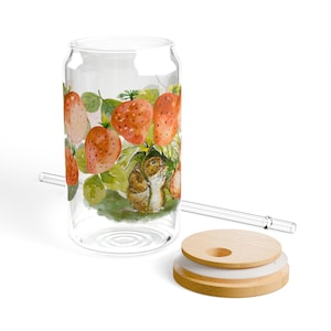Puede incluir: Un vaso transparente en forma de lata con un diseño de fresas y ratón. El vaso presenta una ilustración en acuarela de fresas maduras y un pequeño ratón entre hojas verdes. Incluye una pajita de cristal y una tapa de madera.