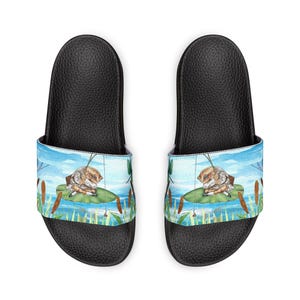May include: Black slide sandals featuring a watercolor illustration of a frog on a lily pad. The design includes a blue sky, water, and reeds. The sandals have a black sole and a printed strap.