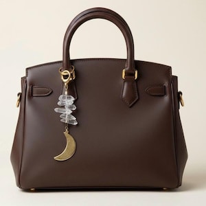 May include: A brown leather handbag with a top handle and gold-colored hardware. A gold-colored crescent moon charm and clear crystal cluster hang from the handle, adding a decorative touch to the bag.
