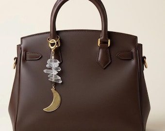 May include: A brown leather handbag with a top handle and gold-colored hardware. A gold-colored crescent moon charm and clear crystal cluster hang from the handle, adding a decorative touch to the bag.