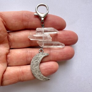 May include: A keychain featuring clear quartz crystals and a silver-toned crescent moon charm. The crystals are arranged in a layered design, suspended from a metal clasp. The moon charm has a textured surface.