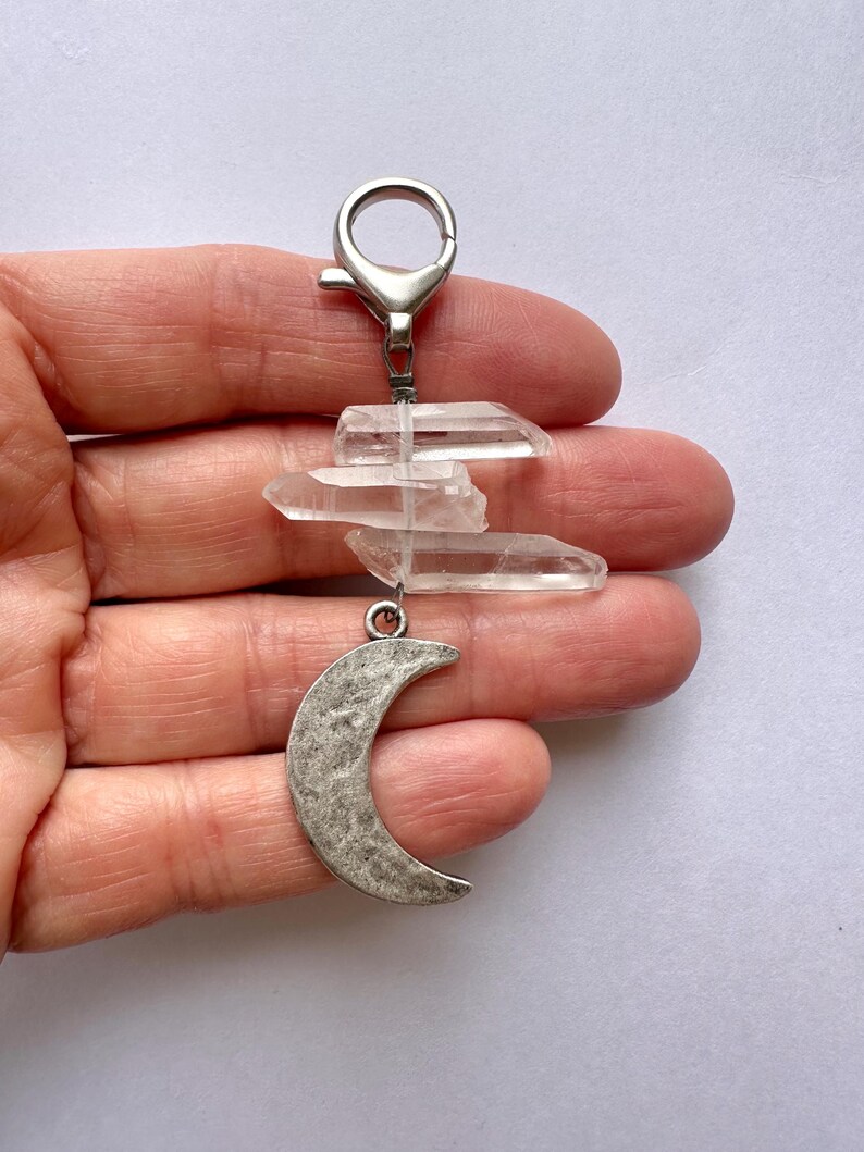 May include: A keychain featuring clear quartz crystals and a silver-toned crescent moon charm. The crystals are arranged in a layered design, suspended from a metal clasp. The moon charm has a textured surface.