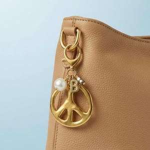 May include: A gold-colored peace sign charm with a pearl and the letter "B" hanging from a gold clasp. The charm is attached to a tan leather handbag. The background is a light blue.