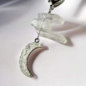 May include: A silver-colored crescent moon charm with a hammered texture hangs below three clear quartz crystals. The crystals are attached to a silver-colored clasp. The charm is set against a white background.