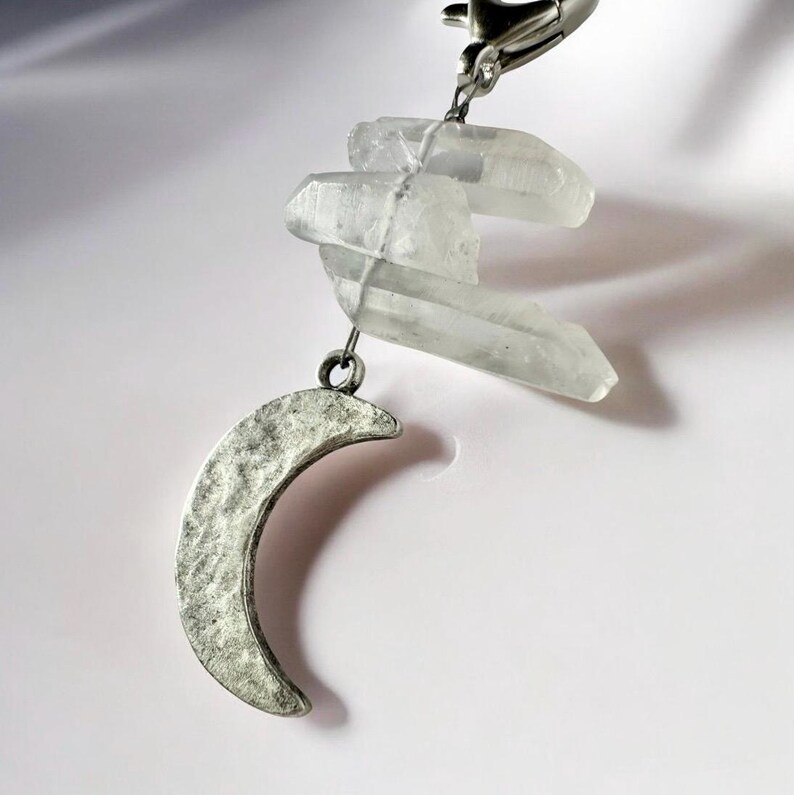 May include: A silver-colored crescent moon charm with a hammered texture hangs below three clear quartz crystals. The crystals are attached to a silver-colored clasp. The charm is set against a white background.