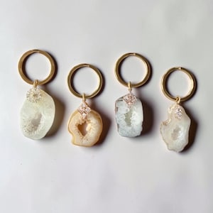 May include: Four gold-toned metal keychains, each featuring a unique, irregular slice of translucent white or pale beige agate stone. Each stone is accented with a small, decorative gold-toned metal charm set with tiny clear crystals at the top.
