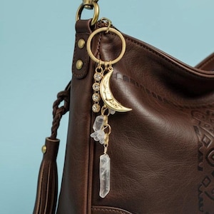 May include: A dark brown leather hobo bag with a braided strap and tassel detail, featuring a gold-toned metal keychain attached to the ring. The keychain includes a crescent moon charm, a vertical row of four clear crystals, and two raw crystal points.
