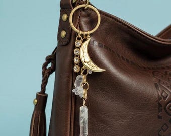 May include: A dark brown leather hobo bag with a braided strap and tassel detail, featuring a gold-toned metal keychain attached to the ring. The keychain includes a crescent moon charm, a vertical row of four clear crystals, and two raw crystal points.