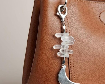 May include: A brown leather handbag with a silver-toned charm. The charm features clear crystal points stacked above a silver crescent moon. The bag has a top handle and a textured surface.