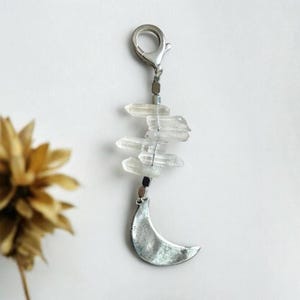 May include: A silver-toned keychain featuring a crescent moon charm and a cluster of clear quartz crystals. The crystals are wire-wrapped and suspended below a clasp. The design has a bohemian or celestial theme.