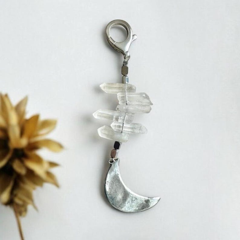 May include: A silver-toned keychain featuring a crescent moon charm and a cluster of clear quartz crystals. The crystals are wire-wrapped and suspended below a clasp. The design has a bohemian or celestial theme.
