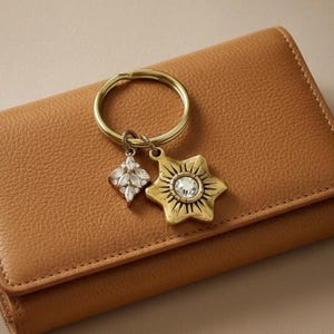 May include: A gold-toned keychain with two charms, one a starburst design with a central crystal and the other a crystal-studded teardrop shape. The keychain rests on a brown leather wallet.