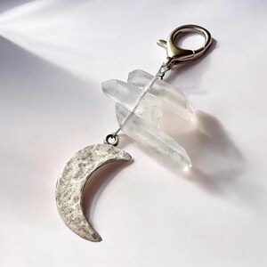 May include: A silver-toned keychain featuring a crescent moon charm and clear quartz crystals. The moon charm has a textured surface, and the crystals are clustered together. The keychain has a silver clasp for easy attachment.