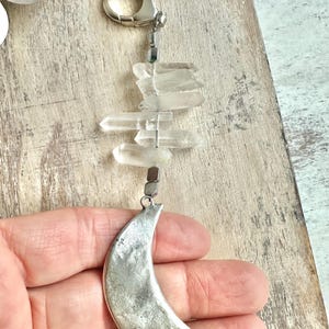 May include: A silver-toned crescent moon charm hangs below clear quartz crystals and a silver clasp. The crystals are arranged vertically, and the moon charm has a textured surface. The charm is held in a hand.