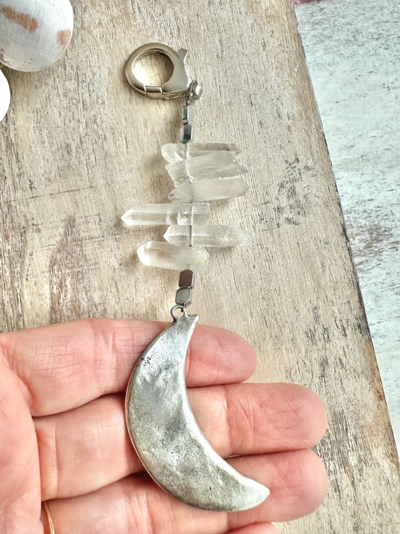 May include: A silver-toned crescent moon charm hangs below clear quartz crystals and a silver clasp. The crystals are arranged vertically, and the moon charm has a textured surface. The charm is held in a hand.