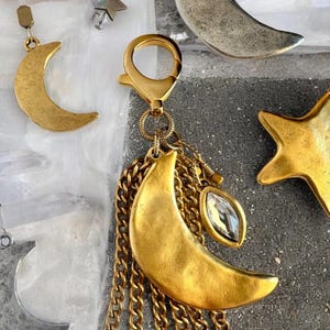 May include: Gold-toned purse charms featuring crescent moons, stars, and chains. One charm has a clear, teardrop-shaped crystal. The charms are attached to a gold-colored clasp. Text on the image reads "Waning Moon and Star Purse Charms".
