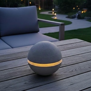 Könnte beinhalten: Eine graue, kugelförmige Außenlampe mit einem horizontalen Band aus warmem Licht. Die Lampe steht auf einem Holztisch, im Hintergrund ein graues Sofa und ein Garten. Die Lampe ist ein dekoratives Beleuchtungselement.