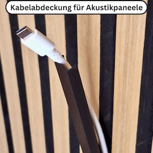 Könnte beinhalten: Nahaufnahme einer schwarzen Kabelabdeckung mit einem weißen USB-C-Kabel. Die Abdeckung ist an einer Holzplatte mit abwechselnd schwarzen und hellbraunen vertikalen Streifen angebracht. Der Text oben lautet "Kabelabdeckung für Akustikpaneele".