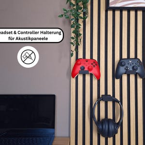 Könnte beinhalten: Eine Wandhalterung für Gaming-Zubehör mit einem roten und einem schwarzen Gamecontroller sowie einem schwarzen Headset. Die Halterung ist an einer Holzpaneele mit schwarzen Streifen befestigt. Der Text "Headset & Controller Halterung für Akustikpaneele" ist sichtbar.