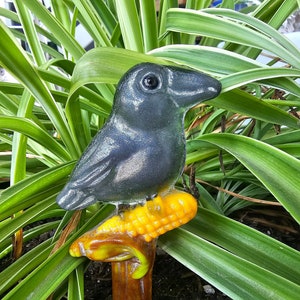 May include: A black crow figurine perched on a yellow corn cob, with a brown wooden base. The figurine is sitting in a pot of green plants.