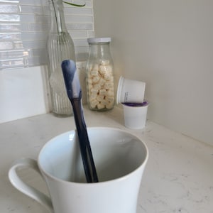 May include: A white ceramic mug with a blue and white stirring stick inside. The mug is on a white countertop with a glass jar filled with white marshmallows, a glass bottle, and a white plastic container.