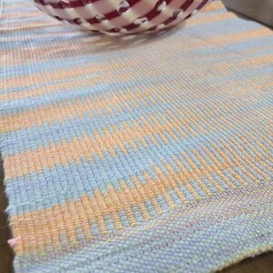 May include: A woven placemat with a blue, pink, and yellow striped pattern. The placemat is made of a soft, textured material and has a slightly frayed edge.