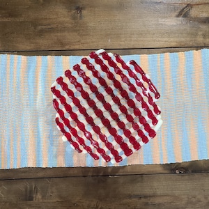 May include: A red and white glass serving dish with a wavy, grid-like pattern. The dish is on a blue and orange striped runner.