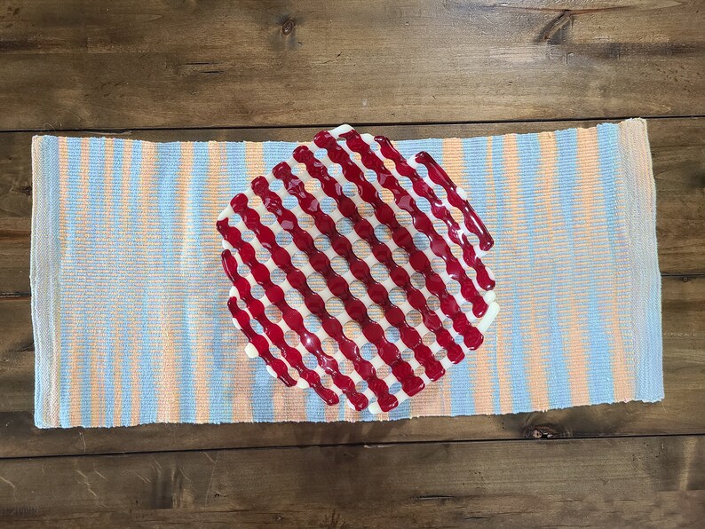 May include: A red and white glass serving dish with a wavy, grid-like pattern. The dish is on a blue and orange striped runner.