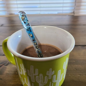 May include: A green mug with a cactus pattern and a brown liquid inside. A blue and white glass spoon is in the mug.