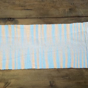 May include: A woven rug with blue and peach stripes. The rug is made of a natural fiber and has a fringed edge.