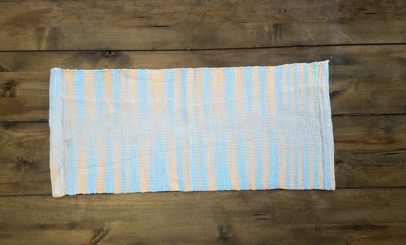 May include: A woven rug with blue and peach stripes. The rug is made of a natural fiber and has a fringed edge.
