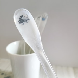 May include: A clear glass spoon with a black and white cartoon character design. The spoon is sitting in a white ceramic mug.