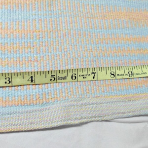 May include: A yellow measuring tape with black markings is laid across a woven fabric with a light blue, pink, and yellow striped pattern. The tape measures 13 inches.
