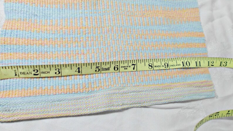 May include: A yellow measuring tape with black markings is laid across a woven fabric with a light blue, pink, and yellow striped pattern. The tape measures 13 inches.