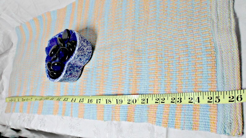 May include: A blue and white woven rug with a measuring tape placed on top. The tape measures 26 inches. A small blue bowl filled with dark blue stones sits on the rug.