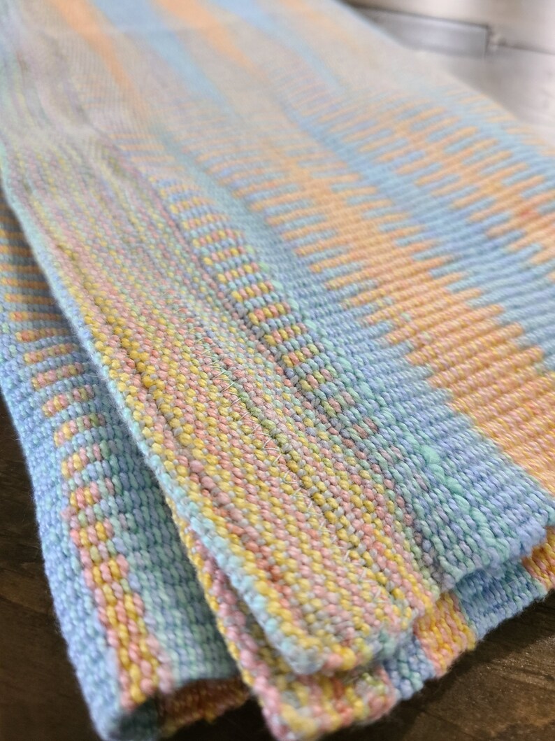 May include: A woven throw blanket with a blue, yellow, and pink striped pattern. The blanket is folded in half and the edges are visible.