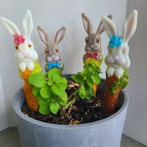 May include: Four ceramic bunny figurines with carrot stems are planted in a gray concrete pot with green leafy plants. The bunnies are white with pink and blue accents and brown with blue accents.