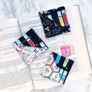 May include: Three black fabric bookmarks with different science-themed patterns. The first bookmark has a pattern of test tubes, molecules, and atoms. The second bookmark has a pattern of mathematical equations and symbols. The third bookmark has a pattern of chemical elements and their symbols.