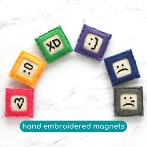 May include: Set of six hand-embroidered felt magnets with different expressions, including a heart, a smiley face, a frown, a sad face, and "XD". The magnets are arranged in a semi-circle on a white marble surface.