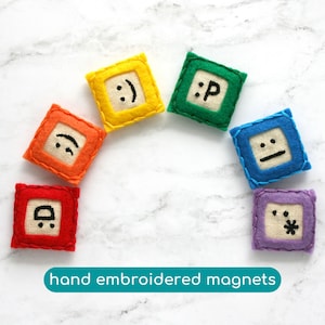 May include: Set of six hand-embroidered felt magnets with different facial expressions. The magnets are in a rainbow colour scheme, with each magnet featuring a different colour: orange, yellow, green, blue, purple, and red.