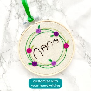 May include: A white embroidery hoop with a green floral wreath and the word "Nana" embroidered in black thread. The wreath is decorated with purple and pink flowers.