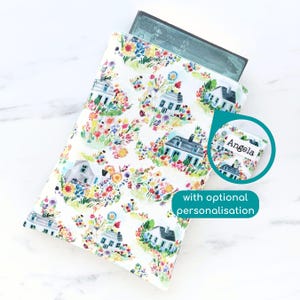 May include: White book sleeve with a colourful floral pattern featuring small houses and the text "Angela" on a white label. The sleeve is designed to fit a standard sized book.