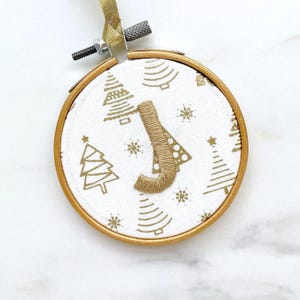 Peut inclure: Ornement de Noël rond dans un cercle à broder. Le tissu blanc est orné de motifs dorés de sapins et de flocons de neige. La lettre "J" est brodée avec du fil beige. Un ruban doré est attaché pour suspendre.