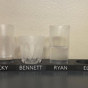 May include: A black wooden serving tray with four circular cutouts for glasses. Three clear glasses are placed in the slots labeled BECKY, BENNETT, and RYAN. The fourth slot labeled ELIZA is empty. The tray features white text for each name.