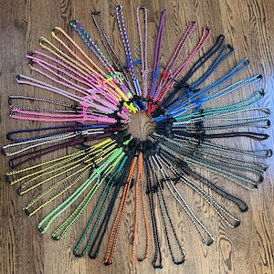 Puede incluir: Una colecci&oacute;n de cordones de paracord coloridos con cierres de metal negro. Los cordones est&aacute;n dispuestos en un c&iacute;rculo sobre una superficie de madera.