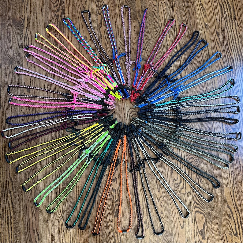 Puede incluir: Una colecci&oacute;n de cordones de paracord coloridos con cierres de metal negro. Los cordones est&aacute;n dispuestos en un c&iacute;rculo sobre una superficie de madera.