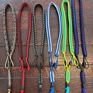 May include: Six paracord lanyards featuring braided designs in various color combinations including camouflage, red and black, olive green and black, white and blue, neon yellow and blue, and purple and black, each with a black plastic buckle.