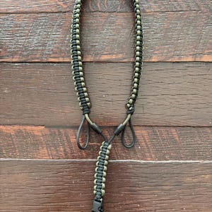 May include: A black and olive green paracord lanyard with a black clasp and loops. The lanyard is made of tightly woven cord and is designed to be worn around the neck. The background is a wooden surface.